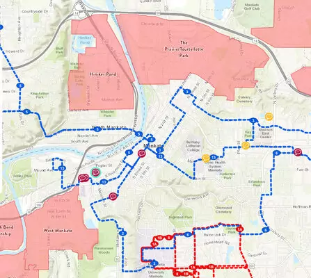 A detailed map showing a city layout with blue, red, and green dotted paths overlaying streets and neighborhoods. Pink shaded areas indicate different zones. Major landmarks and parks are labeled, with a river running through the city.