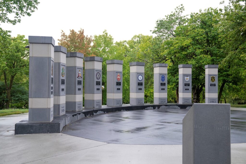 A curved row of tall, gray stone pillars stands on a paved surface at NPHC Plaza, featuring etched military emblems and dates. Lush green trees form a backdrop under a cloudy sky. The area is clean and well-maintained.