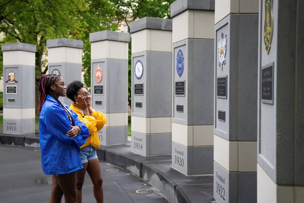 Two people in blue and yellow jackets explore the NPHC Plaza, observing a row of tall, gray pillars adorned with emblems and plaques. The pillars bear dates carved into them, while trees gracefully frame the background.