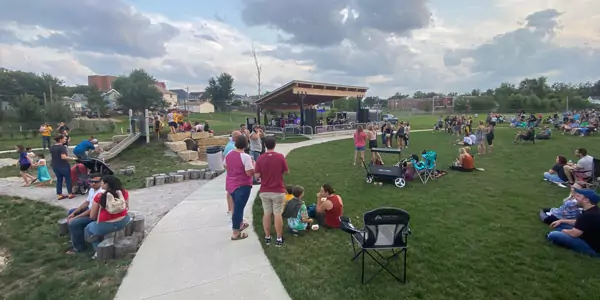 People gather at an outdoor park for a Parks Recreation and Open Space community event with a stage, groups sitting on the grass, walking on paths, and talking. The sky is partly cloudy, and trees border the area.