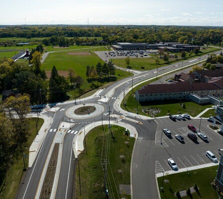 Aerial view of a roundabout connecting multiple roads with crosswalks. Surrounding the roundabout are parking lots, a field, a few buildings, and trees. The image captures a clear day with blue skies and a distant horizon.