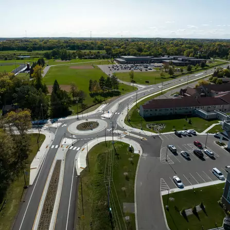 Aerial view of a roundabout connecting multiple roads with crosswalks. Surrounding the roundabout are parking lots, a field, a few buildings, and trees. The image captures a clear day with blue skies and a distant horizon.