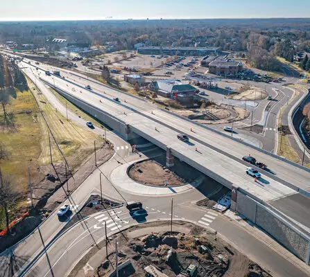 An aerial view of a bridge over an intersection in a suburban area. Cars are driving on the bridge and surrounding roads. Construction equipment and materials are visible below. Buildings and parking lots are in the background under a clear blue sky.