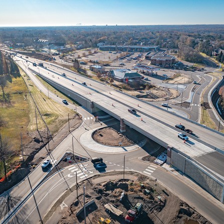 An aerial view of a bridge over an intersection in a suburban area. Cars are driving on the bridge and surrounding roads. Construction equipment and materials are visible below. Buildings and parking lots are in the background under a clear blue sky.