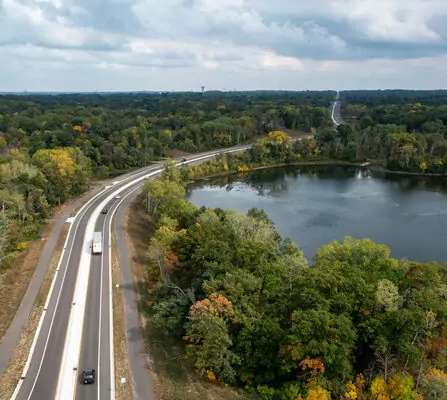 Aerial view of a highway curving alongside a large lake surrounded by dense green forests. Several vehicles are on the road, and the sky is cloudy, casting a soft light over the landscape.