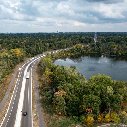 Aerial view of a highway curving alongside a large lake surrounded by dense green forests. Several vehicles are on the road, and the sky is cloudy, casting a soft light over the landscape.