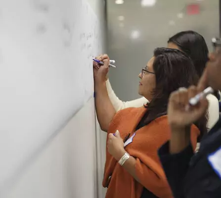 Three people are writing on a large whiteboard. The woman in the foreground is wearing glasses and an orange shawl, writing with a purple marker. The scene suggests a collaborative or educational setting.
