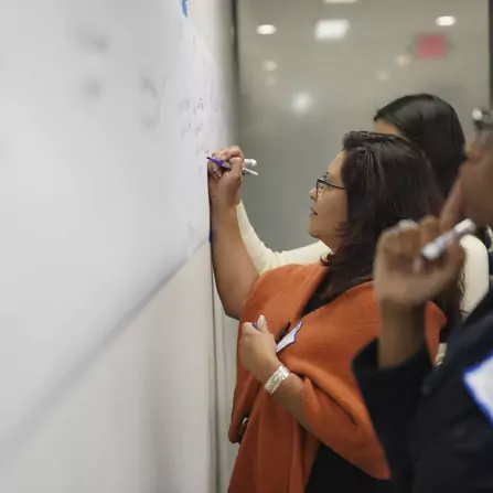 Three people are writing on a large whiteboard. The woman in the foreground is wearing glasses and an orange shawl, writing with a purple marker. The scene suggests a collaborative or educational setting.