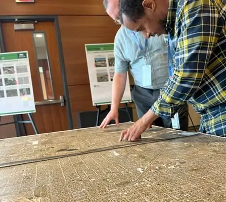 Two people examine a large map on a table in a room with wood-paneled walls. Posters with diagrams are displayed on easels in the background. They are focused, pointing at different sections of the map.