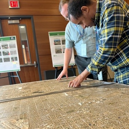 Two people examine a large map on a table in a room with wood-paneled walls. Posters with diagrams are displayed on easels in the background. They are focused, pointing at different sections of the map.