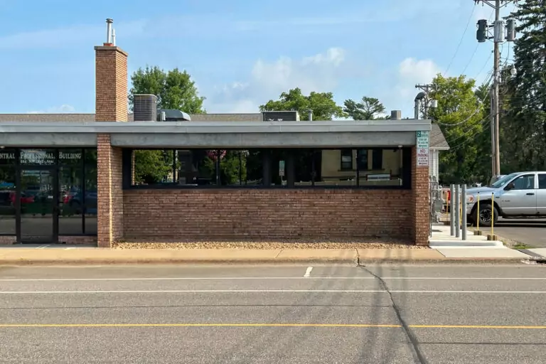 A single-story brick building with a chimney, large windows, and an entrance on the left stands proudly among Bolton & Menk locations. A well-marked parking area is out front, bordered by a road, while trees and utility poles frame the scene in the background.