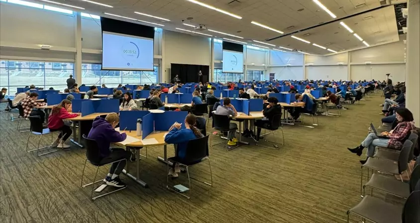 A large room filled with people seated at tables, each separated by blue dividers, exudes the focused atmosphere of a STEAM education event. Participants seem engrossed in an exam or competition, while two large screens display a timer at the front of the room.