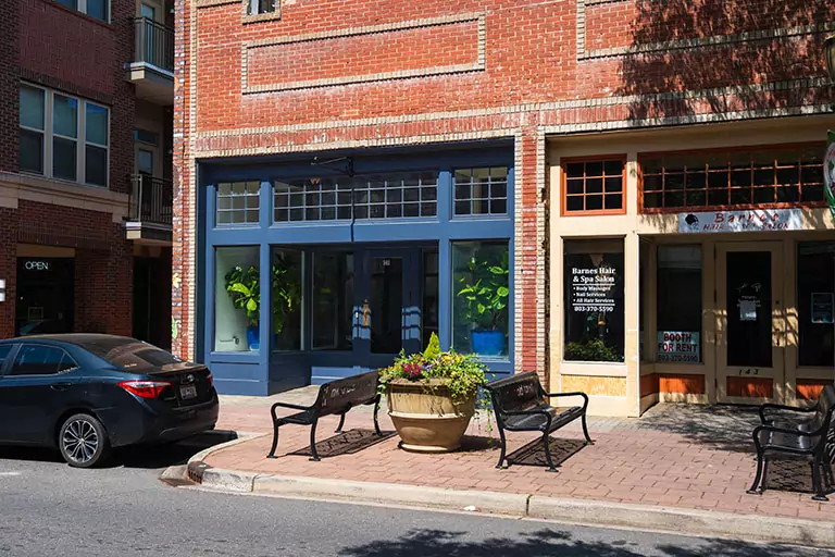 A brick building with large, blue-framed windows houses a tranquil spa salon, reminiscent of Bolton & Menk locations. A car is parked nearby, while two benches sit beside a flower-filled planter. The salons sign highlights its luxurious hair and spa services.