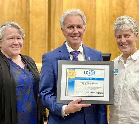 Three people smiling indoors. The person in the center is holding a framed certificate labeled Leadership with a blue and white design. They are standing in front of a wooden wall.