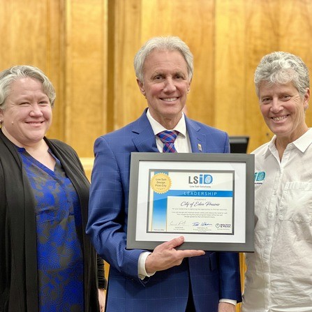 Three people smiling indoors. The person in the center is holding a framed certificate labeled Leadership with a blue and white design. They are standing in front of a wooden wall.