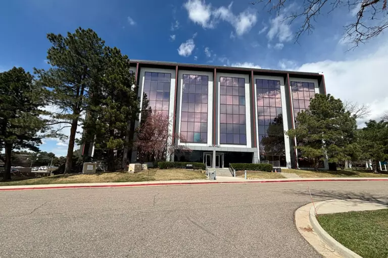 A modern four-story office building with large reflective windows stands among tall pine trees, one of the distinctive Bolton & Menk locations. The partly cloudy sky casts its reflections on the glass as a curved roadway leads to a grassy lawn at the entrance.