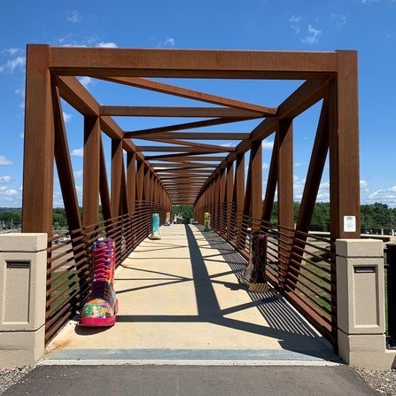 A pedestrian bridge with a rusty metal frame crosses over a landscape. Colorful, oversized cowboy boot statues are placed at intervals along the bridge, and there are trees and a clear blue sky in the background.