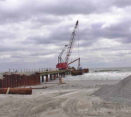 A construction site by the sea with a crane on a pier extending into the water. Steel beams and gravel piles are visible in the foreground, under a cloudy sky. Waves reach the base of the pier.