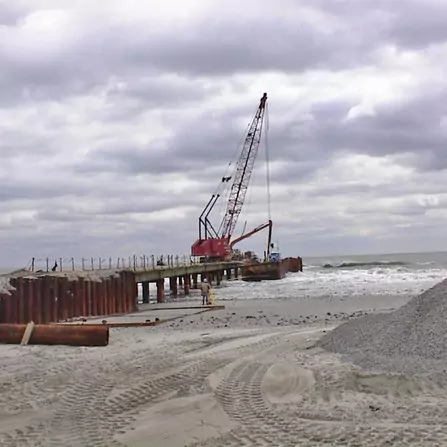 A construction site by the sea with a crane on a pier extending into the water. Steel beams and gravel piles are visible in the foreground, under a cloudy sky. Waves reach the base of the pier.