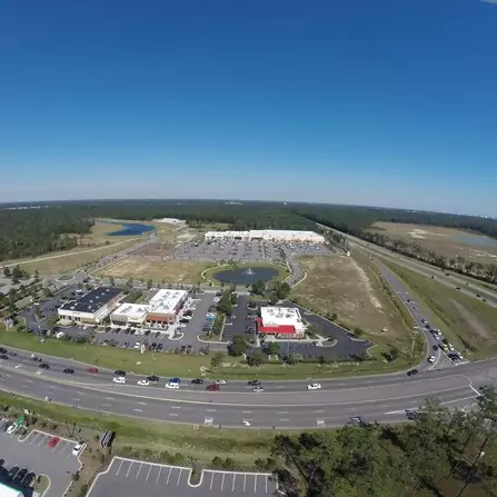 Aerial view of a shopping area with several parking lots, a pond, and a busy road. The background shows a vast expanse of greenery and a clear blue sky.