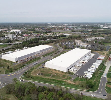 Aerial view of two large warehouse buildings with white roofs, surrounded by parking lots and rows of trucks. Roads and green areas encircle the structures, with more buildings and a distant landscape visible in the background.