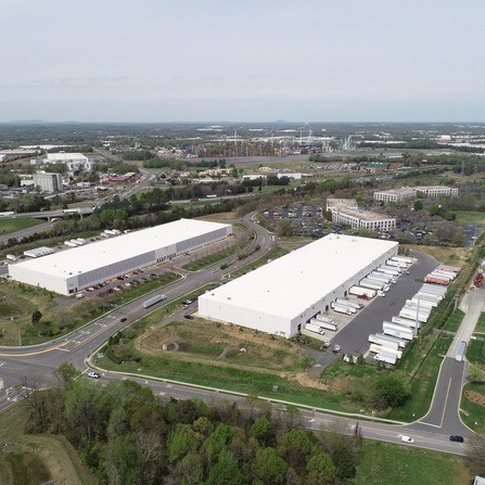 Aerial view of two large warehouse buildings with white roofs, surrounded by parking lots and rows of trucks. Roads and green areas encircle the structures, with more buildings and a distant landscape visible in the background.