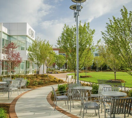 A modern outdoor patio area with metal tables and chairs along a curved pathway. Lush greenery and trees surround the space. A contemporary building with glass windows is in the background. The sky is partly cloudy.