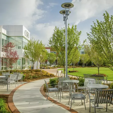A modern outdoor patio area with metal tables and chairs along a curved pathway. Lush greenery and trees surround the space. A contemporary building with glass windows is in the background. The sky is partly cloudy.