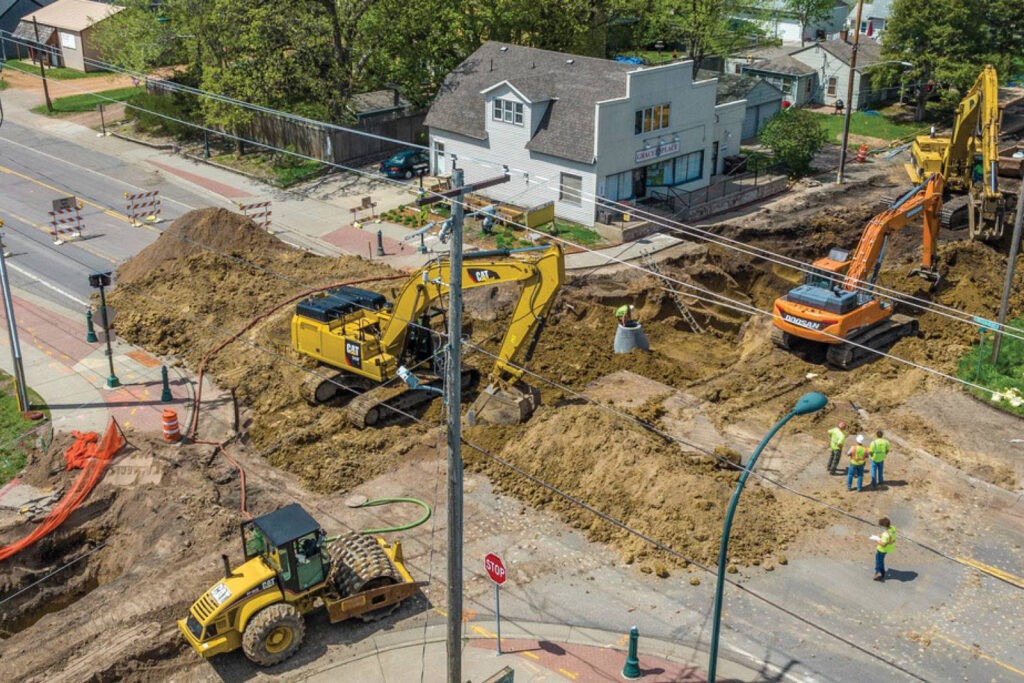 Aerial view of a construction site with several yellow and orange excavators and a bulldozer working on a road. Construction workers in safety gear stand near equipment. Nearby, a two-story house and trees are visible. Traffic cones and barriers are present.