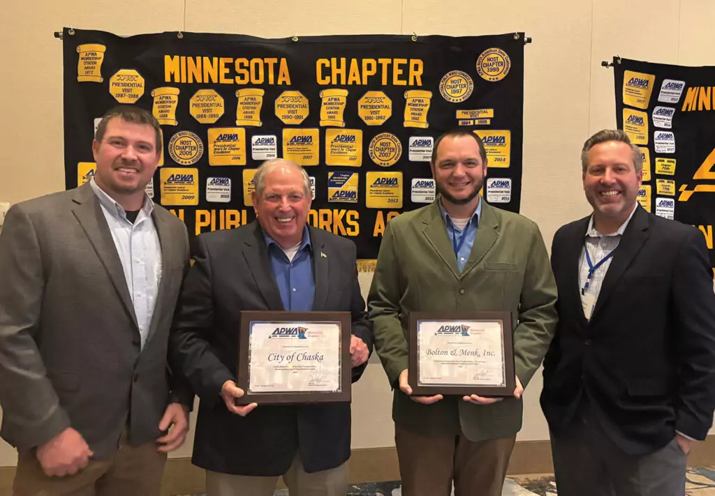 Four men stand smiling, holding plaques awarded during the 2024 APWA-MN Awards in front of a backdrop featuring Minnesota Chapter and various logos. Dressed in business attire, they celebrate their recognition from the public works association.
