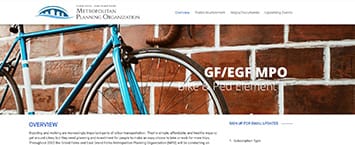 A bicycle is leaned against a red brick wall next to the text GF/EGR MPO Bike & Ped Element, highlighting public projects. The top left corner displays the logo for the Metropolitan Planning Organization.