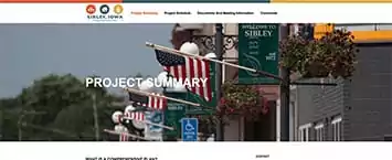 Street view with American flags and hanging plants on lamp posts. Green banners say Welcome to Sibley. The page header reads Project Summary, showcasing one of the towns public projects designed to enhance community pride.