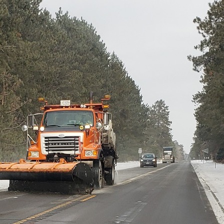 A snow plow on the road with conifers