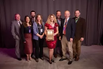 A group of eight people, five men and three women, stand together in formal attire. A woman in the center proudly holds a large framed certificate for Engineering Excellence. They all smile in front of a curtain backdrop on a light-colored floor.