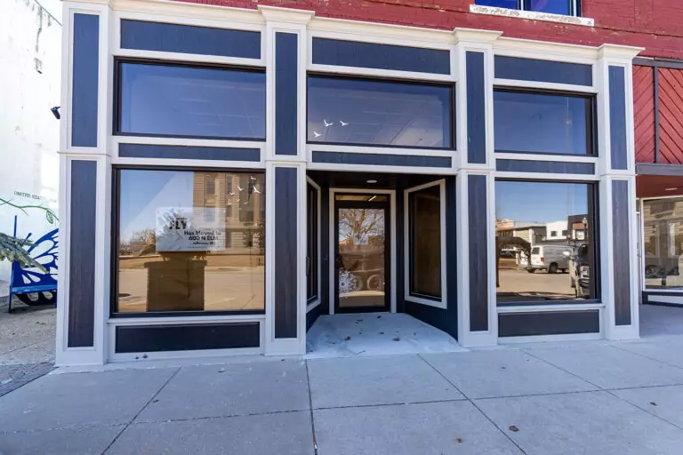A storefront with large glass windows and a black and white facade, reminiscent of many Bolton & Menk locations. The entrance features a recessed door, while a For Lease sign occupies one window. Reflections of buildings and the clear sky dance across the glass surface.