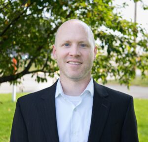 A bald man in a black suit and white shirt stands smiling outdoors, framed by a leafy tree and greenery.