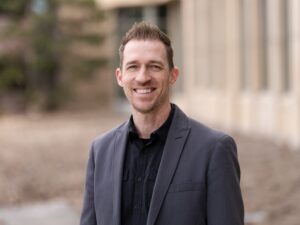 A person with short hair is smiling, wearing a dark blazer and shirt, standing outside a building where the parking lot design offers seamless symmetry amidst the blurred backgrounds.