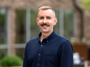 A person with short hair and a mustache is smiling, wearing a dark button-up shirt. The background is blurred, hinting at an outdoor setting where a brick building blends seamlessly with thoughtfully integrated greenery.