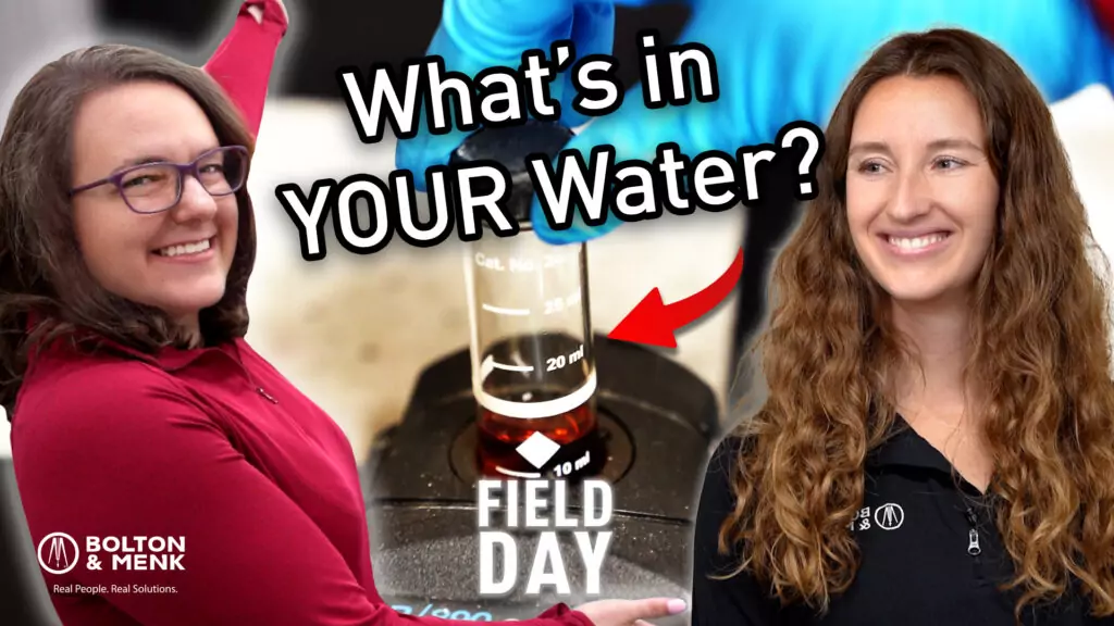 Two people smiling with the text Testing Water: Whats in YOUR Water? overlaid. A red arrow points to a vial filled with liquid marked FIELD DAY. Logos read Bolton & Menk with the tagline Real People. Real Solutions.
