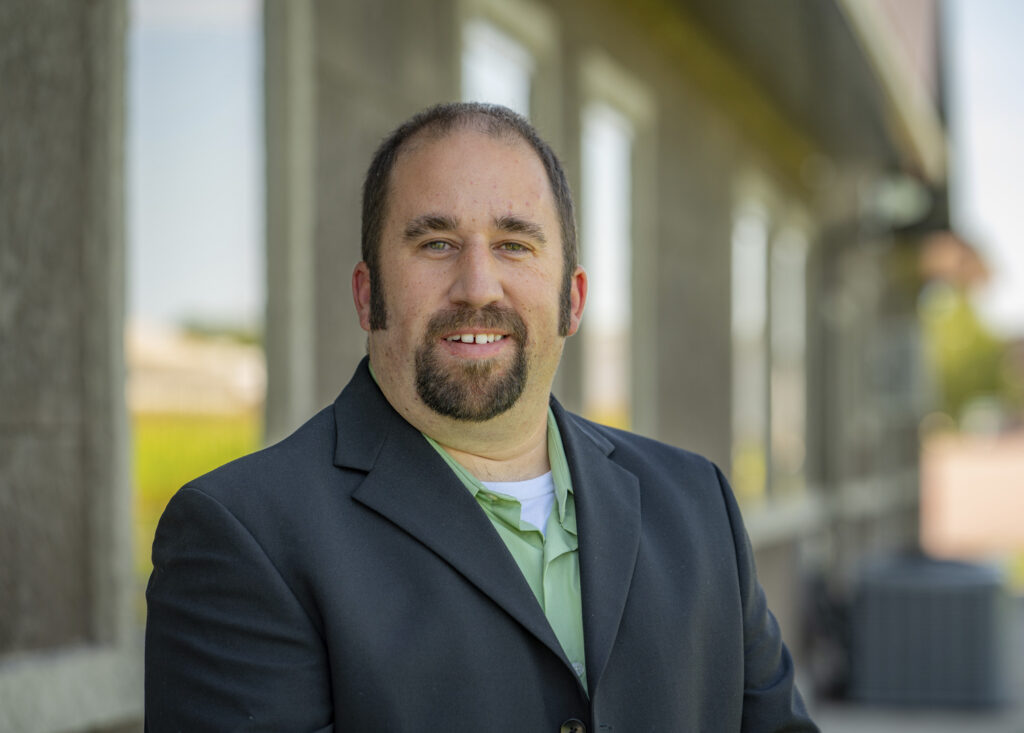 A man with a goatee, wearing a black suit and light green shirt, stands outdoors in front of a building. He is smiling and has a shaved head. The background is blurred, focusing on the man.