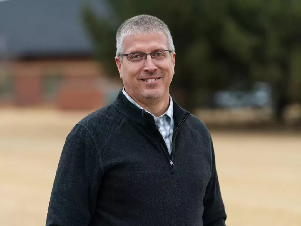 A man with short gray hair and glasses stands outdoors, wearing a dark zip-up sweater over a button-down shirt. He smiles at the camera. The background is blurred and shows trees and a building.