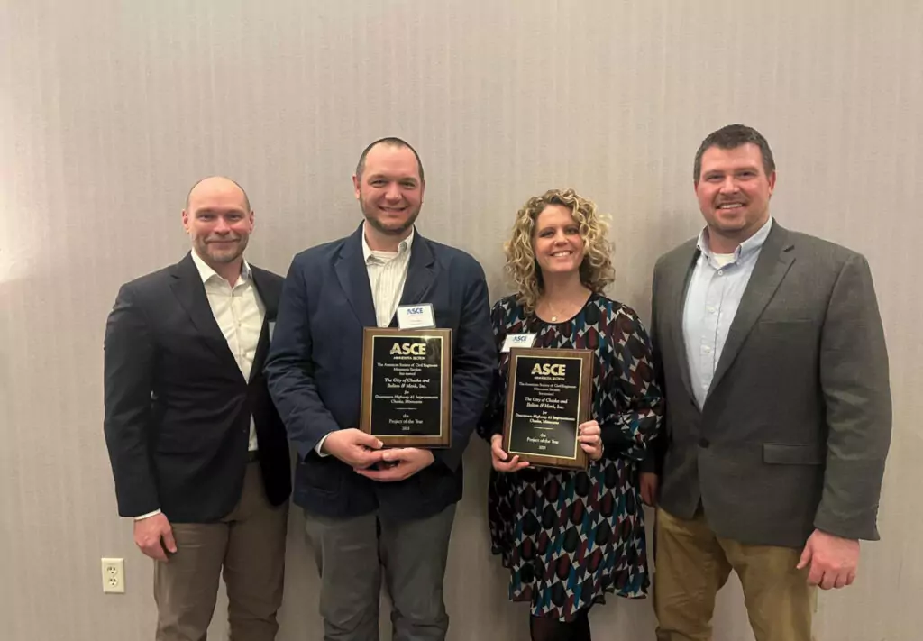 Four people stand in front of a light-colored wall, two holding ASCE award plaques and smiling at the camera. The group appears to be dressed in business or business-casual attire.