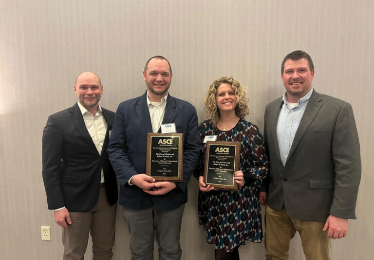 Four people stand in front of a light-colored wall, two holding ASCE award plaques and smiling at the camera. The group appears to be dressed in business or business-casual attire.