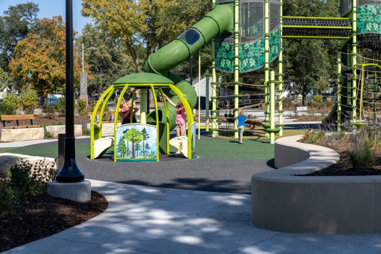 Children play on a green and yellow structure with slides and climbing areas in a park filled with trees, benches, and pathways on a sunny day.