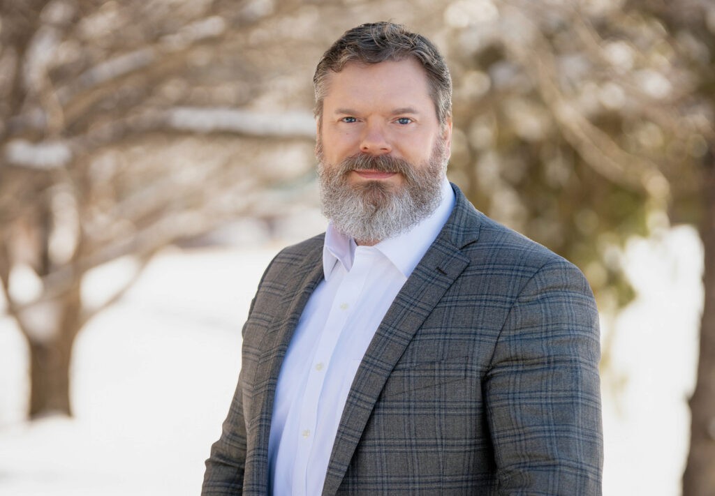 Man with a beard wearing a plaid suit jacket and white shirt stands outdoors. There are blurred trees and a bright, snowy background.