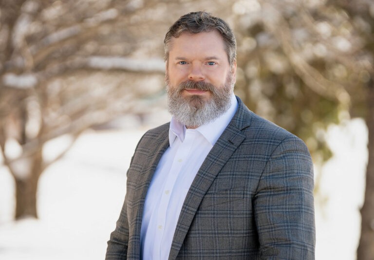 Man with a beard wearing a plaid suit jacket and white shirt stands outdoors. There are blurred trees and a bright, snowy background.