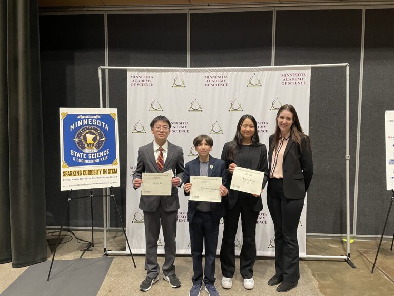 Four students stand together in formal attire, holding certificates at the Minnesota Academy of Science State Science & Engineering Fair. Their smiles celebrate achievements in STEAM Education at this inspiring indoor event.