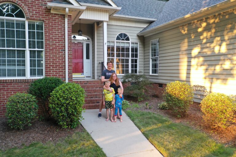 A family of four—two adults and two young children—stand smiling on the walkway in front of a brick house with a red door, surrounded by green bushes and sunlit lawn.
