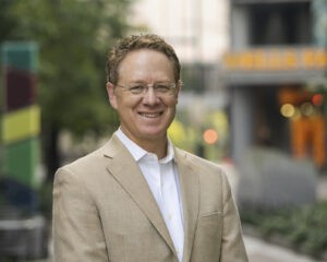 A man in a beige suit jacket and white shirt, smiles while standing outdoors with trees and blurred buildings in the background.
