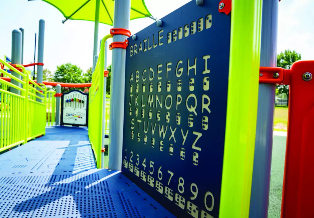 A close-up of a playground panel displaying the Braille alphabet and numbers, with raised braille dots and corresponding letters and numerals. Bright playground structures and green trees are visible in the background.
